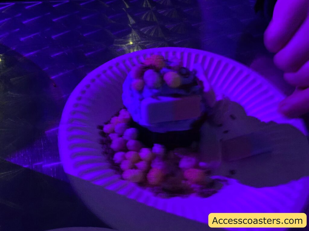 A dessert on a white paper plate, illuminated by purple lighting. The dessert appears to be a cupcake with blue frosting, topped with small round candies in various colors. There are also two rectangular pieces that resemble wafer sticks on top. The plate is placed on a metallic surface with a textured pattern. A hand is visible on the right side of the image. In the bottom right corner, there is a yellow box with the text "Accesscoasters.com".