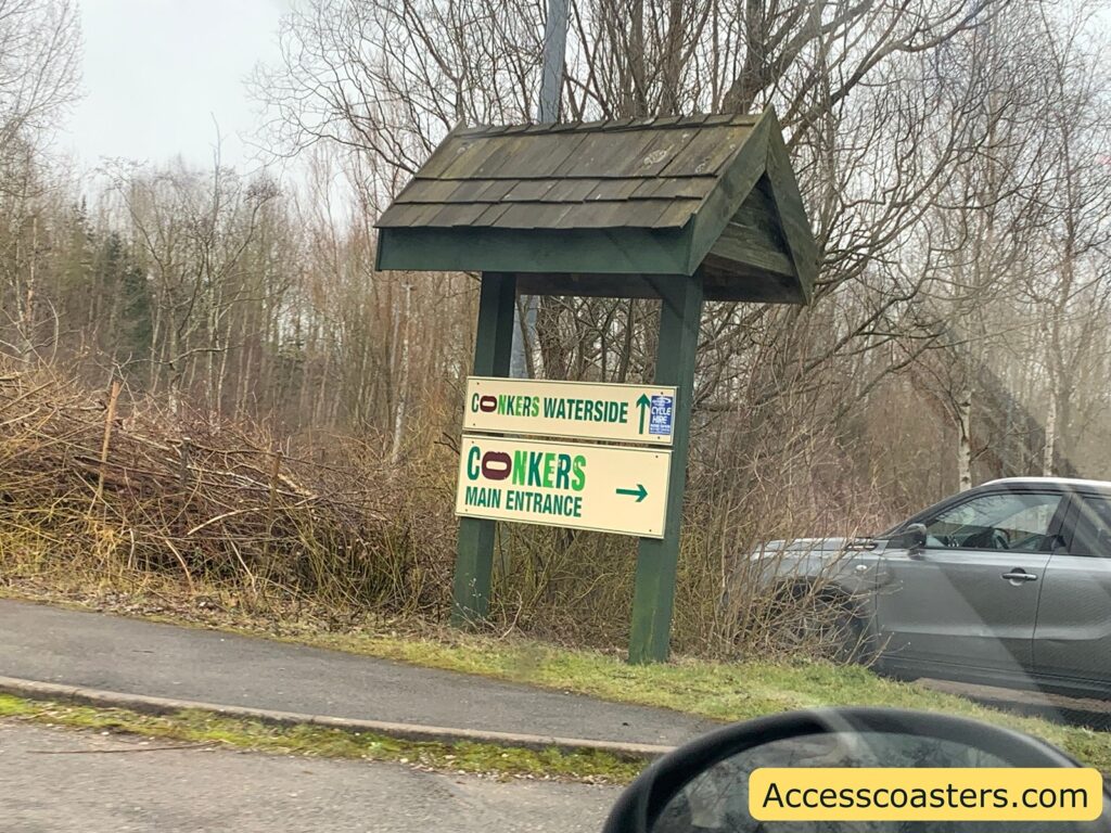 A wooden sign with a roof is situated by the roadside, surrounded by leafless trees and bushes. The sign has two panels. The top panel reads "CONKERS WATERSIDE" with a logo resembling a brown nut in the middle. The bottom panel says "CONKERS MAIN ENTRANCE" with an arrow pointing to the right. There is a small blue sign on the right side of the panel indicating "CYCLE HIRE" with an arrow pointing upwards. A grey car is partially visible on the right side of the image, and the side mirror of another vehicle is visible in the foreground. The website "Accesscoasters.com" is written in yellow at the bottom right corner of the image.