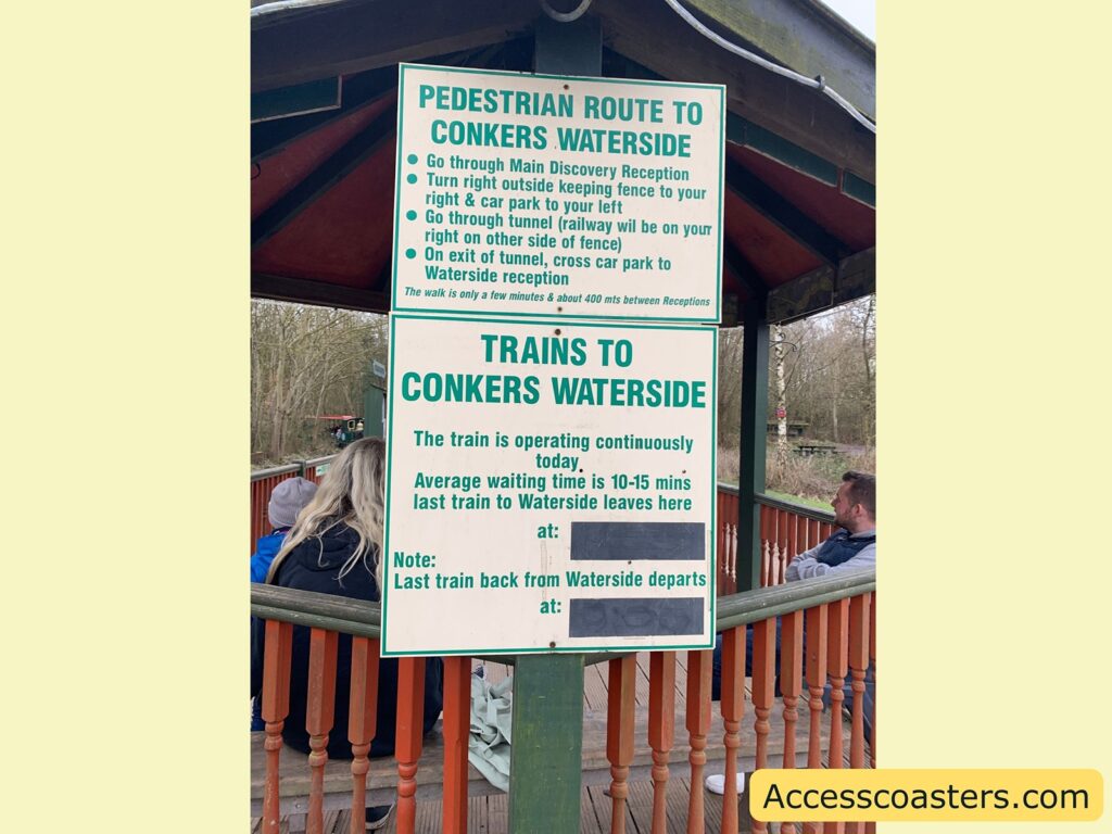 Two signs are displayed at a train station or park area. The top sign provides directions for a pedestrian route to Conkers Waterside:

- Go through Main Discovery Reception
- Turn right outside keeping fence to your right & car park to your left
- Go through tunnel (railway will be on your right on other side of fence)
- On exit of tunnel, cross car park to Waterside reception

It notes that the walk is only a few minutes and about 400 meters between receptions.

The bottom sign provides information about trains to Conkers Waterside:

- The train is operating continuously today.
- Average waiting time is 10-15 minutes.
- Last train to Waterside leaves here at: [blank space]
- Note: Last train back from Waterside departs at: [blank space]

In the background, there are people sitting on benches, and the setting appears to be outdoors with trees visible. The website "Accesscoasters.com" is mentioned at the bottom right corner.
