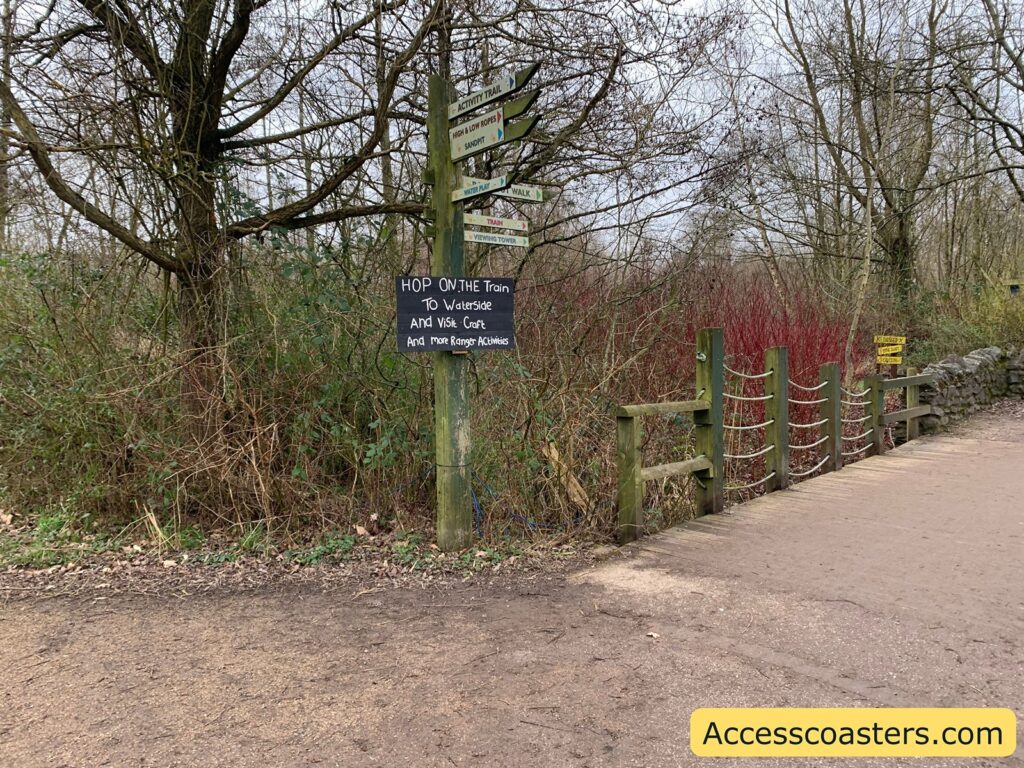 A dirt path runs through a wooded area with leafless trees and bushes. On the left side, there is a wooden post with several directional signs pointing in different directions. One of the signs reads, "HOP ON THE Train TO Waterside And Visit Craft And More Ranger Activities." To the right, a wooden fence with rope sections runs alongside the path. In the bottom right corner, there is a yellow box with the text "Accesscoasters.com."