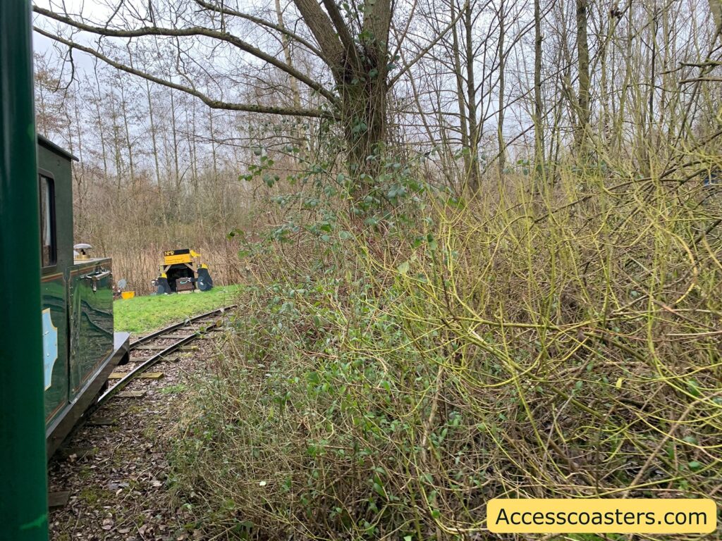 A narrow-gauge railway track curves through a wooded area with bare trees and dense undergrowth. On the left, part of a green train is visible, with a window and a portion of its side. In the background, a small yellow maintenance vehicle is parked on the grass near the tracks. The sky is overcast, and the overall atmosphere is that of a quiet, natural setting. In the bottom right corner, there is a yellow banner with the text "Accesscoasters.com".