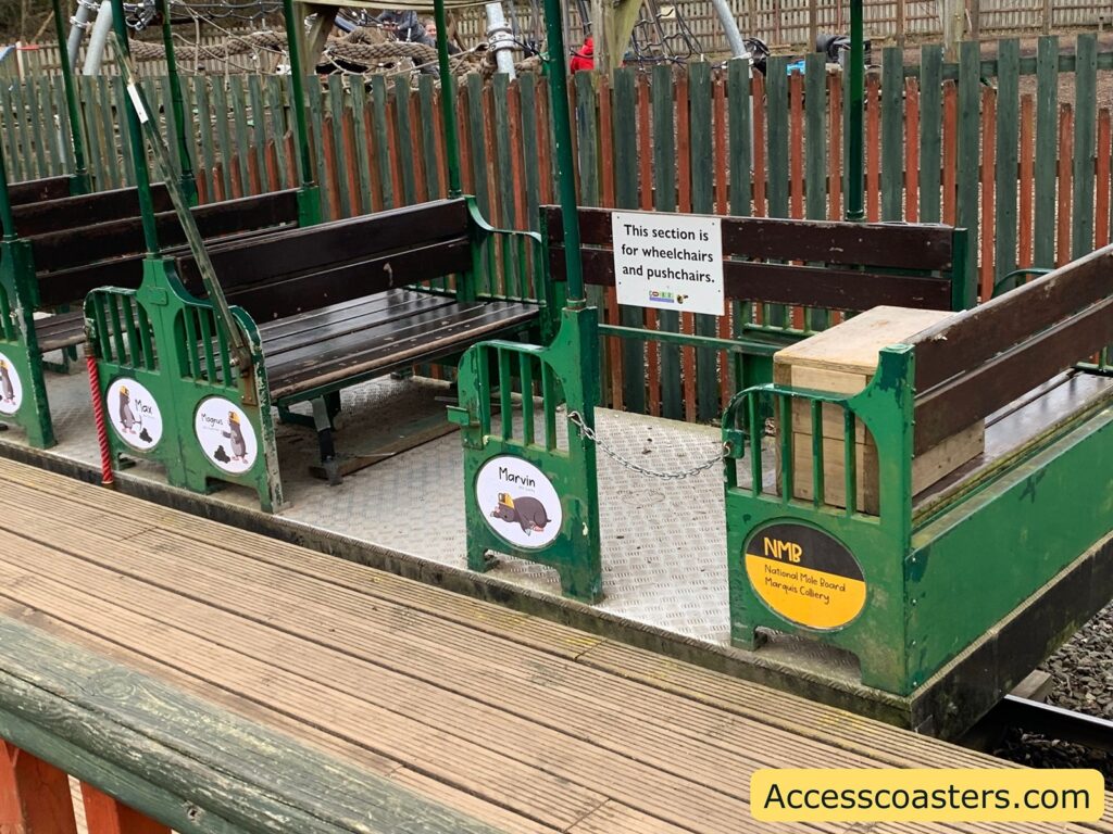A section of a small train or tram with wooden benches and green metal frames. There is a sign on one of the benches that reads, "This section is for wheelchairs and pushchairs." The train is on a track with a wooden platform beside it. The side of the train has circular signs with cartoon images of moles and names like "Max," "Mogul," and "Marvin." Another sign reads "NMB National Mole Board Marquis Colliery." In the bottom right corner, there is a yellow rectangle with the text "Accesscoasters.com." The background shows a wooden fence and some playground equipment.
