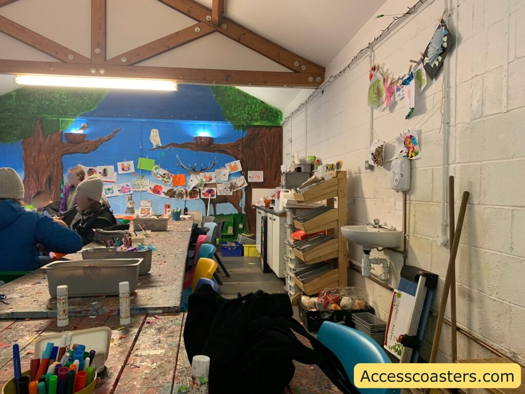 A colorful and creative art room with a long table covered in art supplies like markers and paint bottles. Several people are seated at the table, wearing winter hats and jackets, suggesting a cold environment. The walls are decorated with children's artwork, including drawings and paintings, some of which are hung on a string with clothespins. The back wall features a mural of trees with an owl perched on a branch. There are shelves filled with more art supplies and a small sink on the right side. The room has a high ceiling with wooden beams. In the bottom right corner, there is a yellow label with the text "Accesscoasters.com".