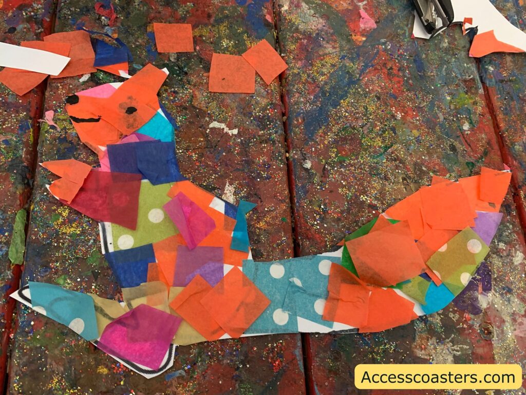 A colorful collage of a squirrel made from various pieces of tissue paper in different colors and patterns, including orange, blue, pink, red, and green with white polka dots. The squirrel is set against a vibrant, multicolored background covered in glitter. In the bottom right corner, there is a yellow label with the text "Accesscoasters.com".