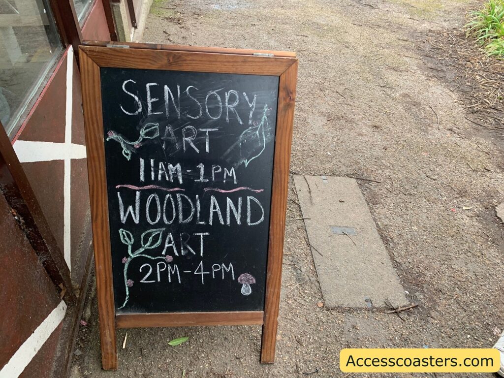 A wooden-framed chalkboard sign stands on a gravel path. The sign reads:

"SENSORY ART
11AM - 1PM
WOODLAND ART
2PM - 4PM"

The text is written in white chalk, with decorative elements like leaves and mushrooms drawn in colored chalk. To the right of the sign, there is a patch of grass. In the bottom right corner of the image, there is a yellow rectangle with the text "Accesscoasters.com" written in black.