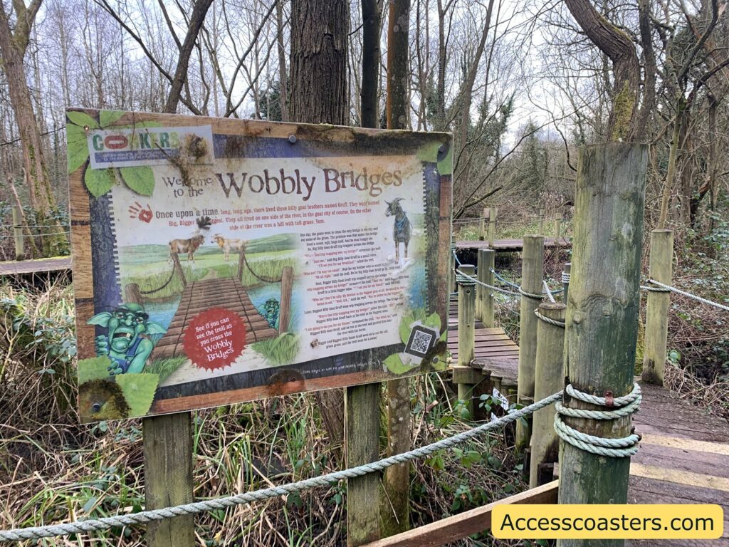 A wooden signboard is displayed in a wooded area with leafless trees and a wooden pathway. The sign reads "Welcome to the Wobbly Bridges" and features a story about three billy goat brothers named Gruff. The story is accompanied by illustrations of a wooden bridge, a troll, and a goat. The sign invites visitors to see if they can spot the troll as they cross the Wobbly Bridges. There is a QR code at the bottom right corner of the sign. The website "Accesscoasters.com" is mentioned on a yellow banner at the bottom right of the image.