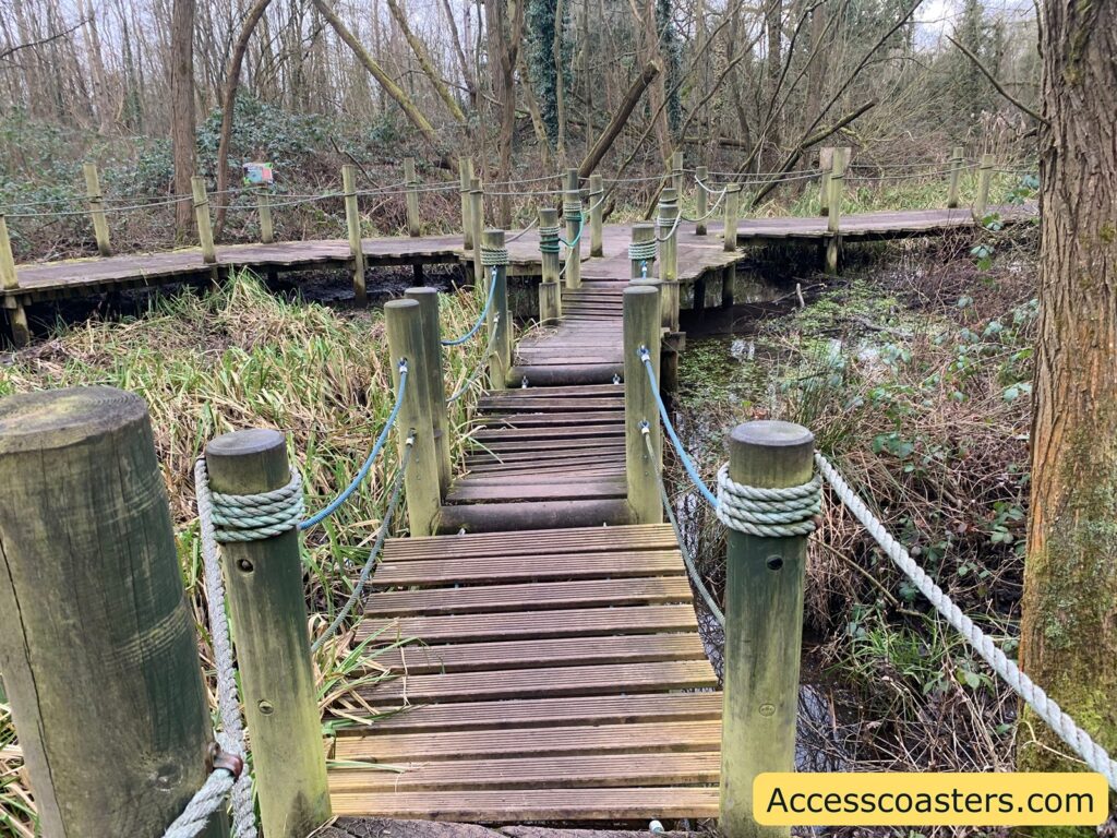 A wooden boardwalk path winds through a marshy, wooded area. The path is elevated on wooden posts and has rope railings on either side for safety. The surrounding area is filled with tall grasses and sparse trees, with some undergrowth visible. The atmosphere is natural and serene, suggesting a nature trail or park setting. In the bottom right corner, there is a yellow banner with the text "Accesscoasters.com".