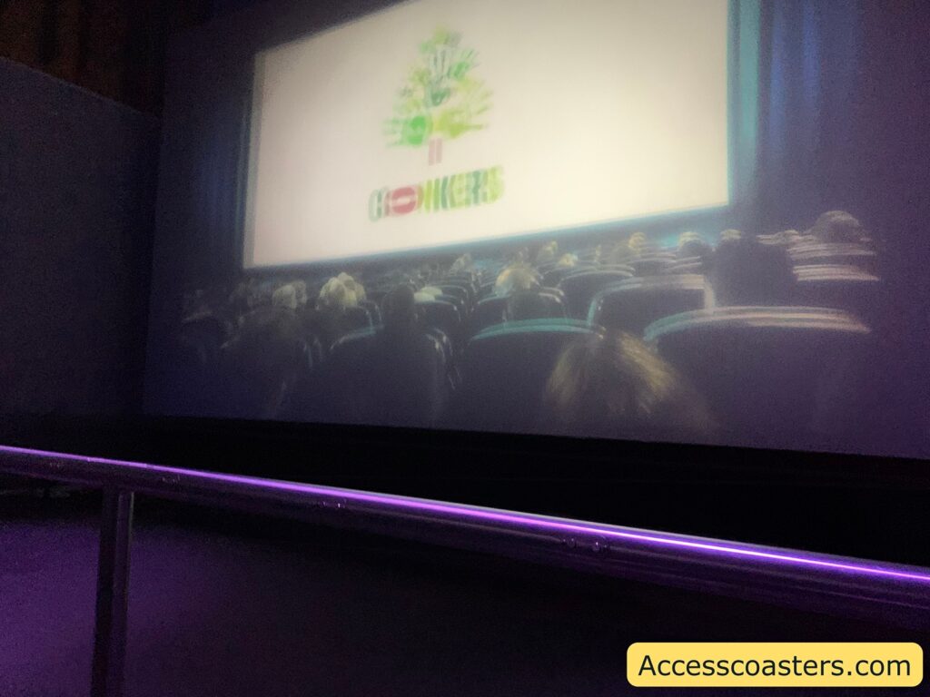 A dimly lit movie theater with rows of seats filled with people. The large screen at the front displays a logo or image that is not clearly visible due to the lighting and angle. The theater has a railing in front of the seats, and the atmosphere is dark, typical of a cinema setting. In the bottom right corner, there is a yellow box with the text "Accesscoasters.com".