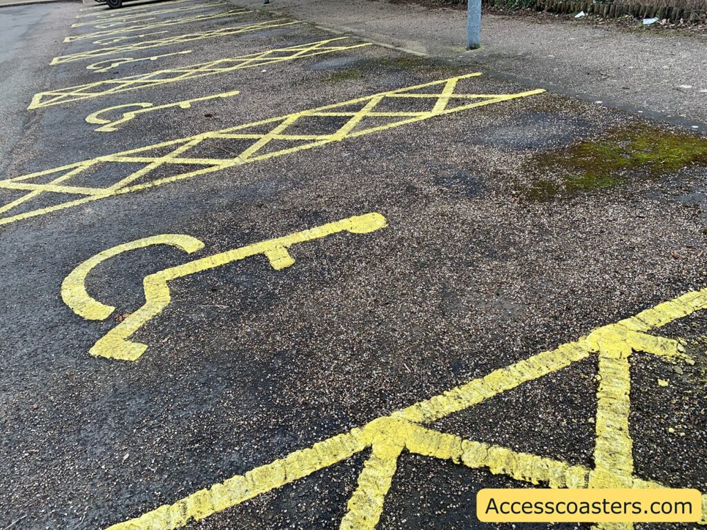 Parking spaces with yellow markings on a rough asphalt surface. The spaces are designated for disabled parking, indicated by the wheelchair symbol painted in yellow. There are also yellow cross-hatched lines next to the parking spaces, which are typically used to indicate no parking zones to allow space for wheelchair access. In the bottom right corner, there is a yellow label with the text "Accesscoasters.com".