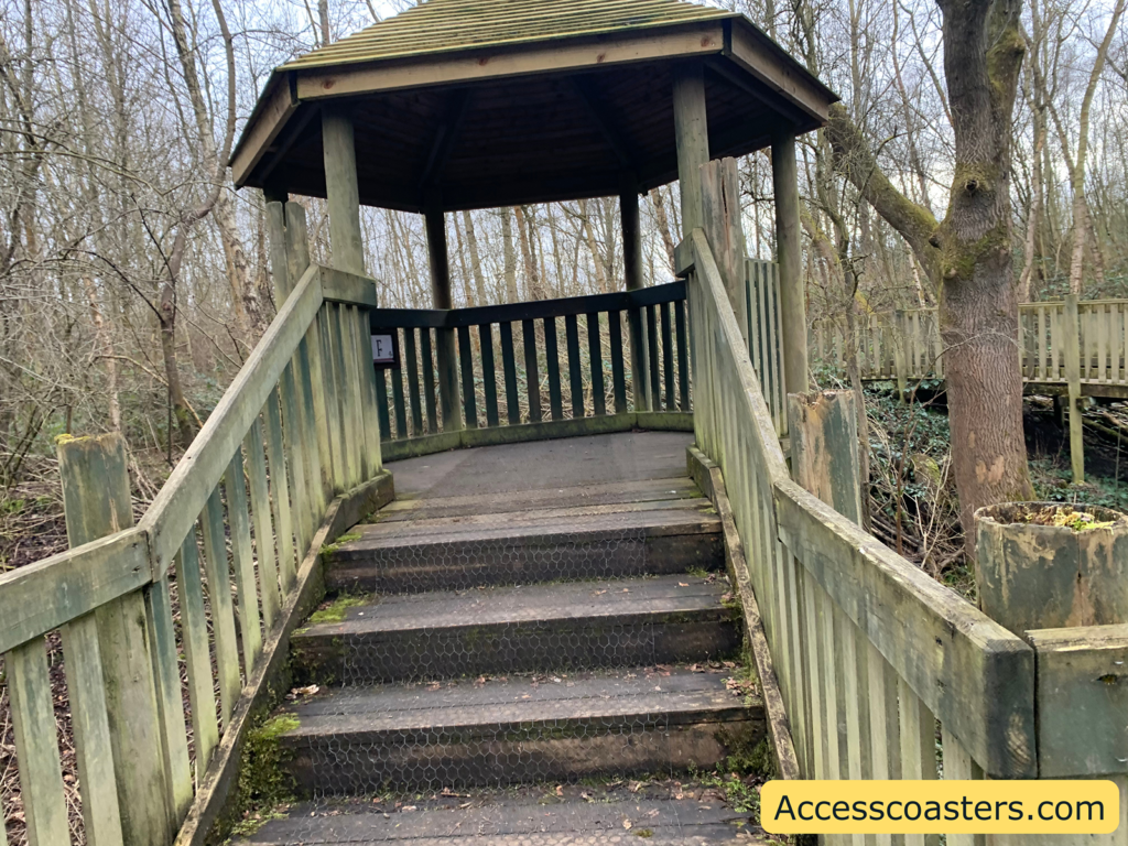 Wooden gazebo structure in a forested area, accessible by a short flight of steps with railings on both sides. The gazebo has a hexagonal roof and is surrounded by bare trees, indicating a winter or early spring setting. The steps and railings show signs of weathering, with some moss growth. A wooden walkway extends from the gazebo into the forest. In the bottom right corner, there is a yellow label with the text "Accesscoasters.com".