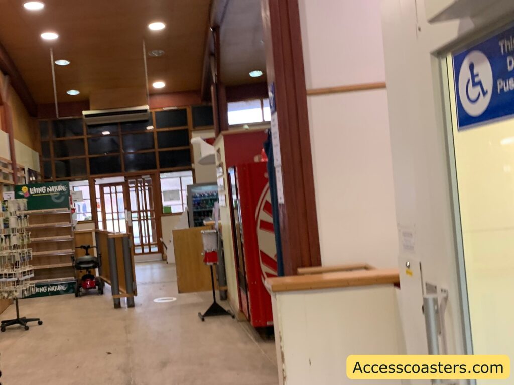 Interior of a store or public space with a high ceiling and wooden accents. On the left, there are shelves with a sign that reads "LIVING NATURE." In the middle, there is a mobility scooter parked near a counter. To the right, there is a vending machine with a red and white design. Near the vending machine, a sign with a wheelchair symbol indicates accessibility. The entrance at the back has large glass doors. The image has a watermark in the bottom right corner that reads "Accesscoasters.com."