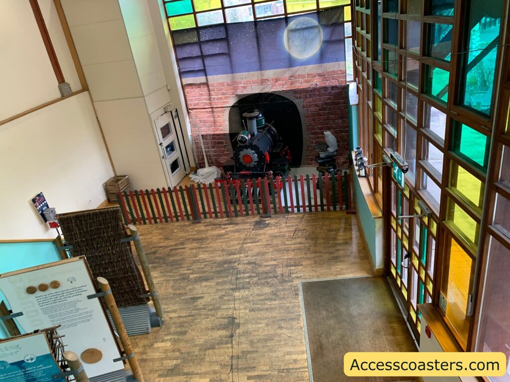A room with wooden flooring and large windows with colorful stained glass panels. In the corner, there is a small, decorative train engine behind a red and green picket fence. The walls are painted white, and there is a mural of a brick wall with a moonlit sky above the train. On the left side, there are informational panels with text and images, supported by wooden posts. Near the bottom right corner, there is a yellow box with the text "Accesscoasters.com".