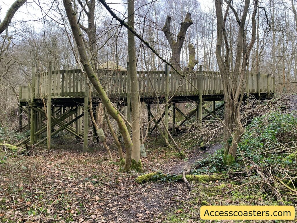 A wooden elevated platform or walkway is situated in a forested area. The structure is supported by wooden beams and has a railing around it. The surrounding area is densely wooded with leafless trees, indicating a winter or early spring setting. The ground is covered with fallen leaves, branches, and some green undergrowth. In the bottom right corner, there is a yellow box with the text "Accesscoasters.com".