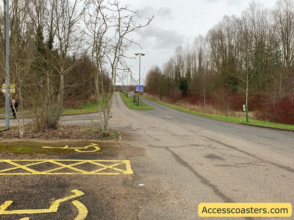 A rural road flanked by leafless trees and bushes on both sides. The sky is overcast, giving a grayish tone to the scene. In the foreground, there is a parking area with yellow-painted markings for disabled parking spaces. The road extends into the distance, with a few signs visible along its sides. In the bottom right corner, there is a yellow label with the text "Accesscoasters.com".