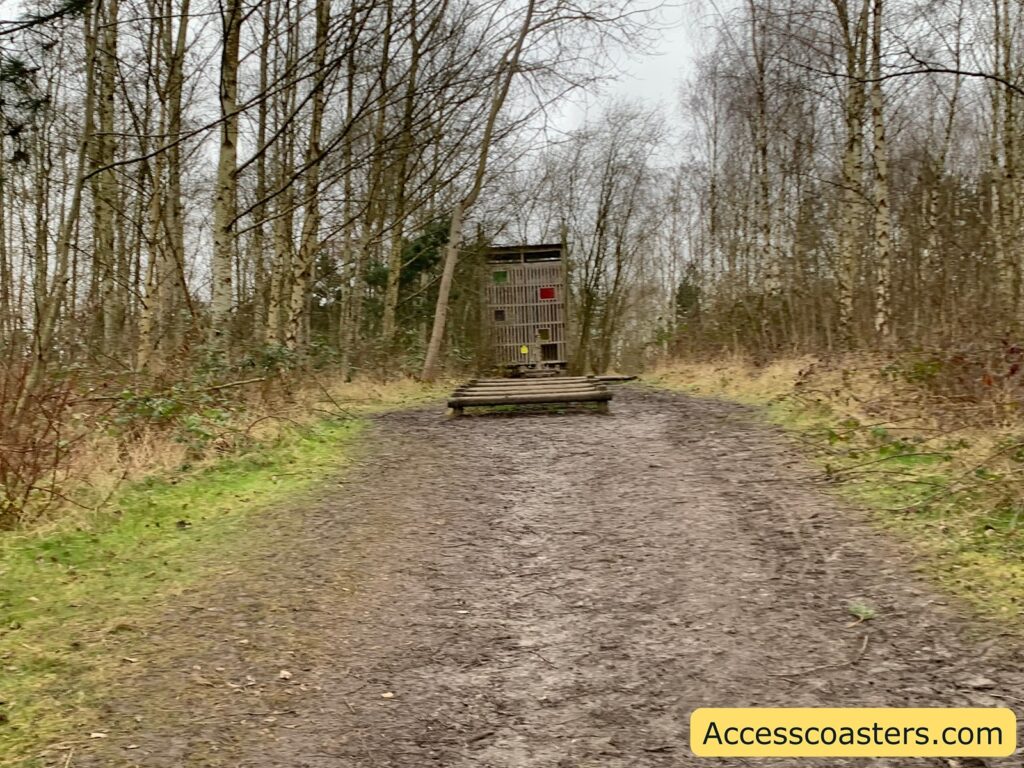A muddy path leads through a forest of bare trees, suggesting a winter or early spring setting. In the distance, there is a wooden structure resembling a small observation tower or lookout, with a ladder leading up to it. The ground is bordered by patches of green grass and some low shrubs. The sky is overcast, adding a gray tone to the scene. In the bottom right corner, there is a yellow box with the text "Accesscoasters.com".