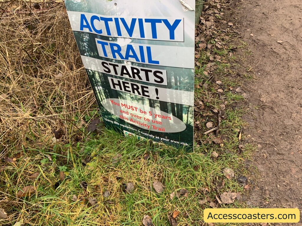 A sign next to a dirt path and grass reads: "ACTIVITY TRAIL STARTS HERE!" Below that, in red text, it says: "You MUST be 5 years and over to use this Activity Trail." At the bottom, there's a cautionary note: "Using this equipment is entirely at your own risk." In the bottom right corner, there's a yellow label with the text "Accesscoasters.com". The background of the sign features an image of a forest.