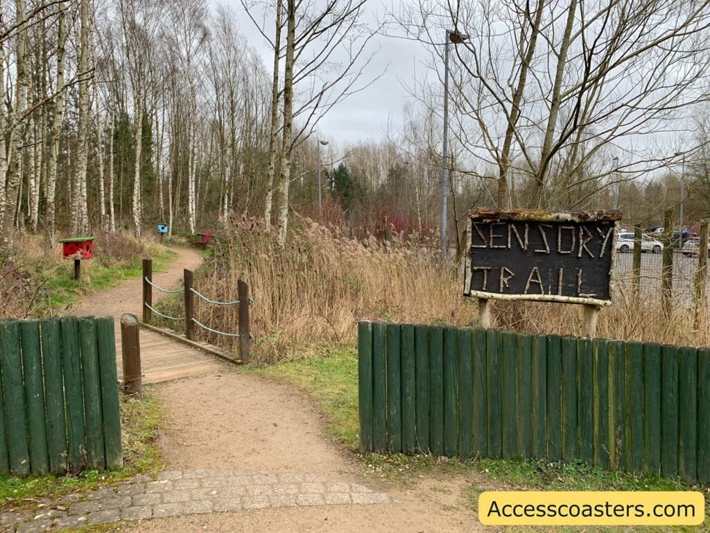 A dirt path leads into a wooded area with tall, leafless trees. The path is bordered by a low, dark green wooden fence. A small wooden bridge with rope railings crosses over a section of the path. To the right, there is a rustic wooden sign with the words "SENSORY TRAIL" written on it. In the background, there are colorful structures along the path, possibly for interactive or sensory experiences. The sky is overcast, and there are parked cars visible in the distance. In the bottom right corner, there is a yellow box with the text "Accesscoasters.com".