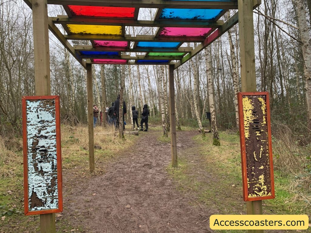 A wooden structure with a series of colored glass panels on top, including red, yellow, pink, blue, and green. The structure is situated on a dirt path surrounded by a forest of leafless trees. On either side of the path, there are two vertical panels with textured surfaces; one is light blue and the other is yellow. In the background, a group of people is gathered, some wearing winter clothing. In the bottom right corner, there is a yellow rectangle with the text "Accesscoasters.com