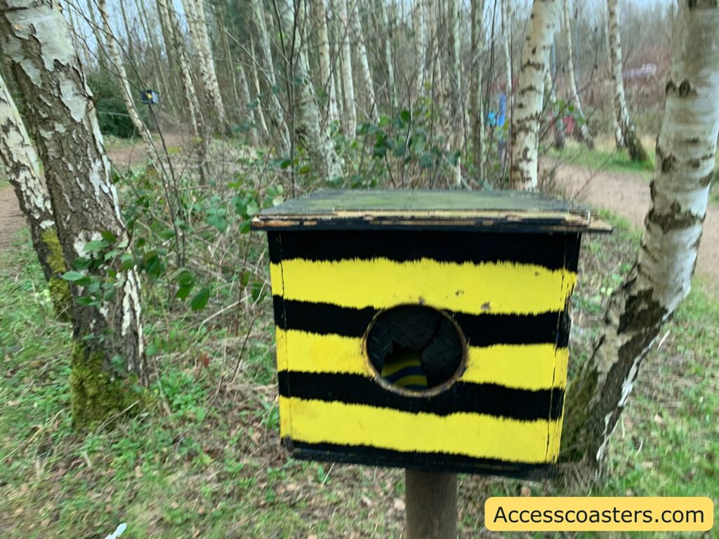 A wooden box painted with alternating black and yellow horizontal stripes, resembling a bee's pattern. It has a circular hole in the center, and inside the hole, there appears to be a tire or rubber object. The box is mounted on a wooden post among a grove of birch trees with white bark. The ground is covered with grass and some low shrubs. In the bottom right corner, there is a watermark with the text "Accesscoasters.com".