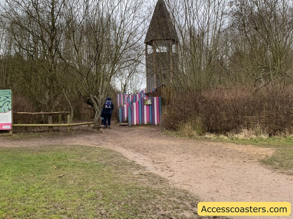 A person wearing a blue jacket and a backpack is walking towards a colorful entrance made of vertical wooden planks painted in various pastel colors. The path is surrounded by bare trees and bushes, suggesting a winter or early spring setting. In the background, there is a tall wooden structure with a pointed roof, resembling a tower. A sign with information is visible on the left side, and the ground is a mix of grass and dirt. The bottom right corner of the image has the text "Accesscoasters.com" on a yellow background.
