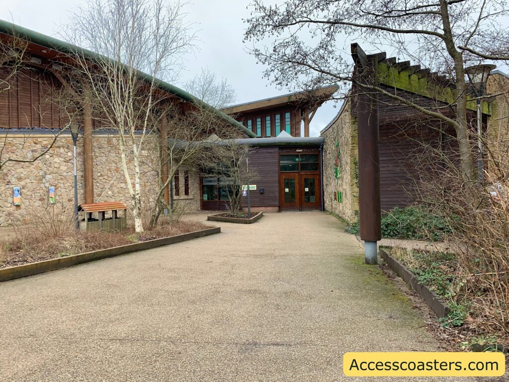 A building with a rustic design, featuring stone and wooden elements. The entrance has large wooden doors and is surrounded by windows with wooden frames. The roof has a green, slightly curved design. There are leafless trees and shrubs in the foreground, with a paved pathway leading to the entrance. The building is set in a natural environment, with some greenery visible. In the bottom right corner, there is a yellow rectangle with the text "Accesscoasters.com".