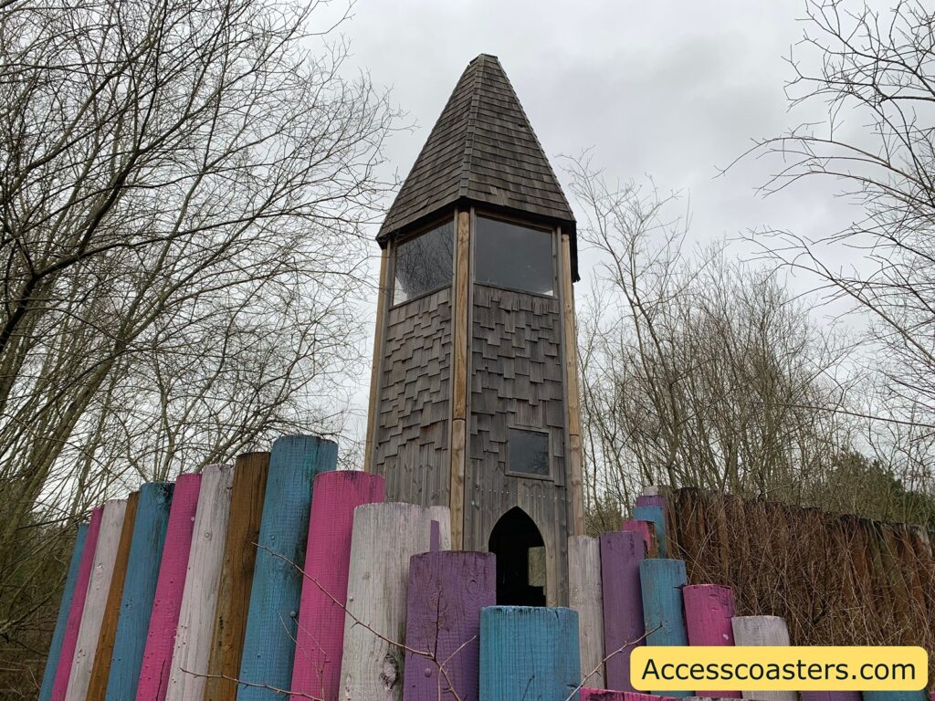 A tall, wooden tower with a pointed roof stands amidst bare trees. The tower has a rustic appearance with wooden shingles and a small arched entrance. Surrounding the base of the tower is a colorful fence made of vertical wooden planks painted in shades of blue, pink, purple, and white. The sky is overcast, giving the scene a muted, wintry feel. In the bottom right corner, there is a yellow banner with the text "Accesscoasters.com".