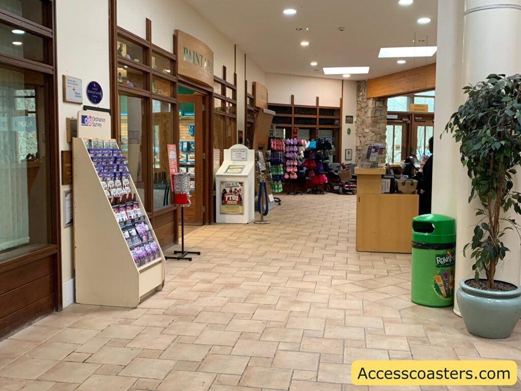 A hallway with a tiled floor and wooden-framed glass doors and windows on the left. There is a display stand with Ordnance Survey maps and brochures. A sign above the door reads "PAINT A POT." In the background, there are shelves with various colorful items, possibly clothing or accessories. A wooden counter is on the right, with a person standing behind it. A green trash bin with "Rowntree's" branding is next to a potted plant on the right. The image has a watermark in the bottom right corner that reads "Accesscoasters.com."