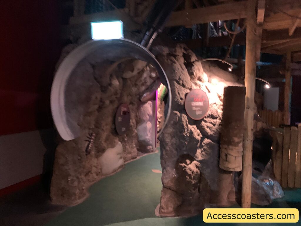 Dimly lit indoor setting with a rocky cave-like structure. There's a circular tunnel entrance on the left side, resembling a large pipe. The walls of the structure have a rough, stone-like texture with some artificial insects attached to it. A circular sign is mounted on the wall, partially illuminated by overhead lights. The floor is covered with green carpeting. In the bottom right corner, there's a yellow label with the text "Accesscoasters.com".