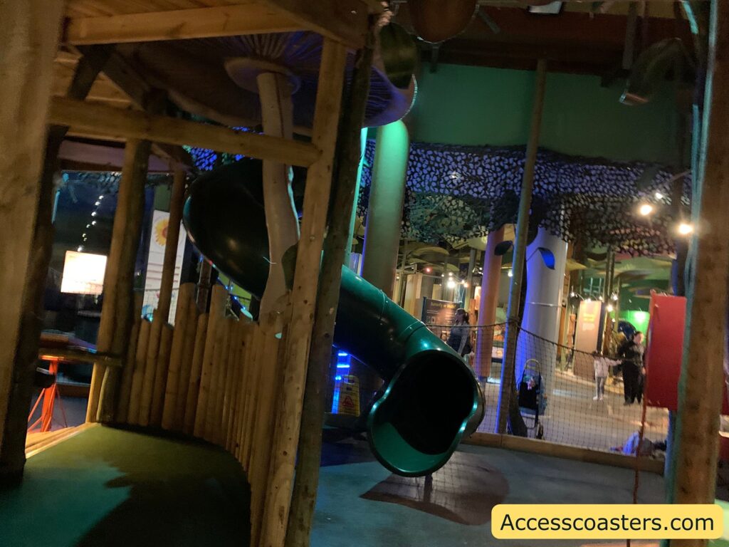 Indoor playground area with a large green spiral slide. The surroundings have a jungle or forest theme, with wooden structures and decorative elements resembling trees and foliage. There are people in the background, including children playing. The lighting is dim, with colorful lights adding to the playful atmosphere. In the bottom right corner, there is a watermark that reads "Accesscoasters.com".