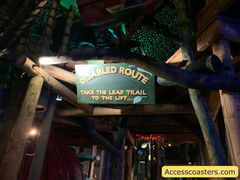 A wooden structure with a sign hanging from it. The sign reads: "DISABLED ROUTE TAKE THE LEAF TRAIL TO THE LIFT..." The background is dimly lit with a greenish hue, and there are wooden beams and netting visible. In the bottom right corner, there is a yellow label with the text "Accesscoasters.com".
