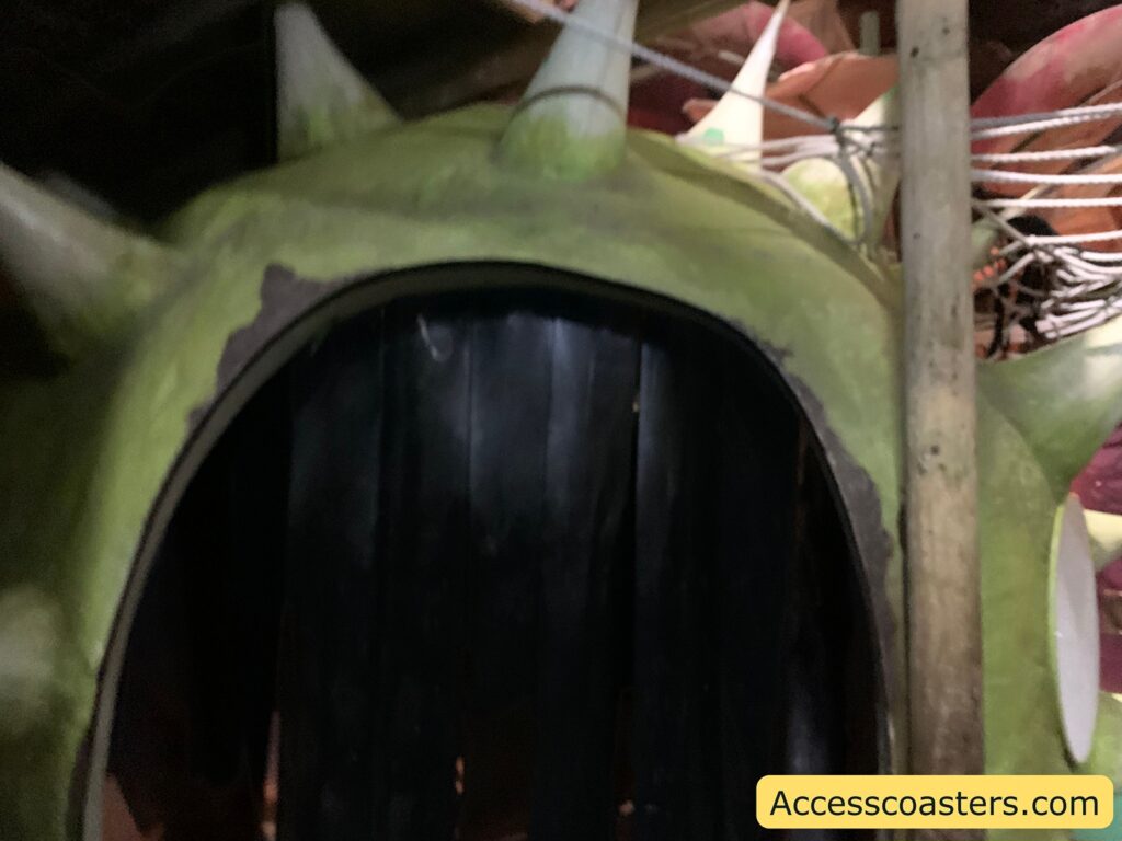 A large, green, spiky structure with an opening that resembles a mouth or entrance. The structure has a rough texture and appears to be part of a themed environment, possibly a ride or attraction. There are ropes and wooden poles around it, adding to the theme. The bottom right corner has the text "Accesscoasters.com."