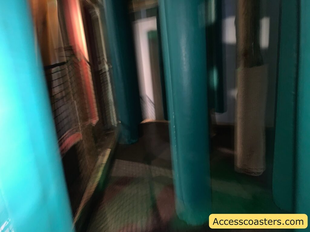 Blurred image of a space with several large, vertical, cylindrical structures in a teal color. There is a netted barrier on the left side. The environment appears to be indoors, possibly part of an amusement or play area. In the bottom right corner, there is a yellow rectangle with the text "Accesscoasters.com".