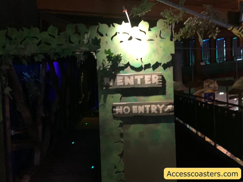 A dimly lit entrance with a jungle or forest theme. The entrance is adorned with artificial leaves and branches. There are two wooden signs on a green wall with the words "ENTER" and "NO ENTRY" painted in white. The area beyond the entrance is dark, with a few small green lights on the floor. In the bottom right corner, there is a yellow box with the text "Accesscoasters.com".