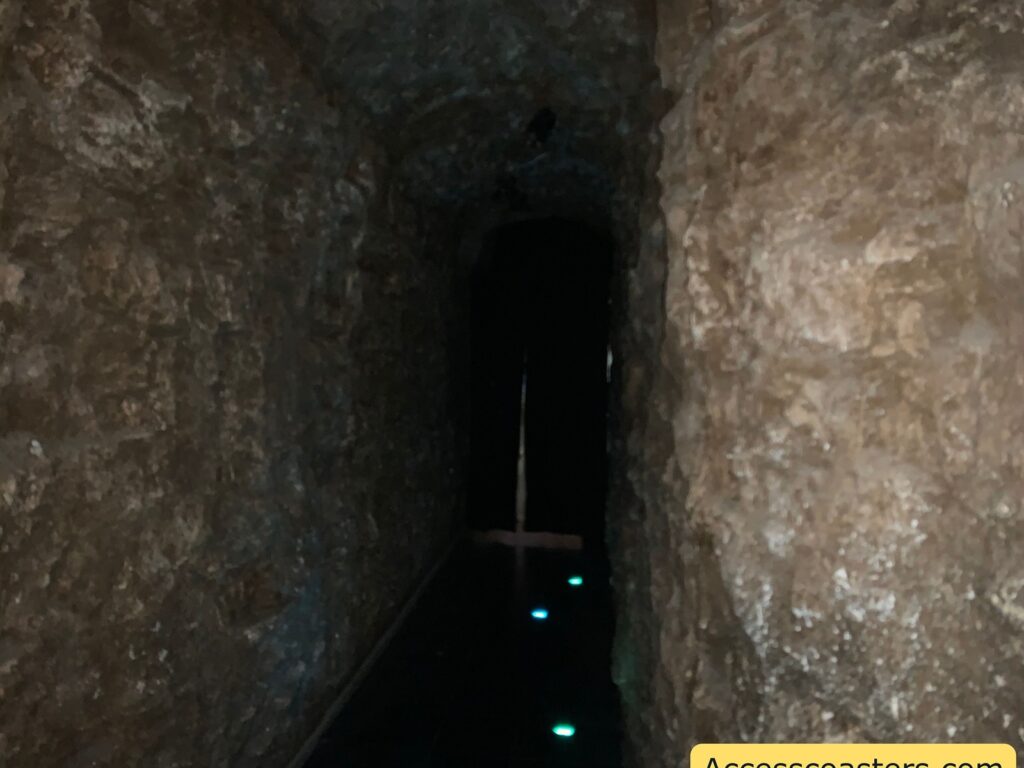 A narrow, dimly lit tunnel with rough, rocky walls. The tunnel is dark, with a few small, faint blue lights on the ground leading into the darkness. The walls appear to be made of natural stone, giving the impression of a cave or underground passage. In the lower right corner, there is a partial view of a yellow object with text that reads "Accesscoasters.com."
