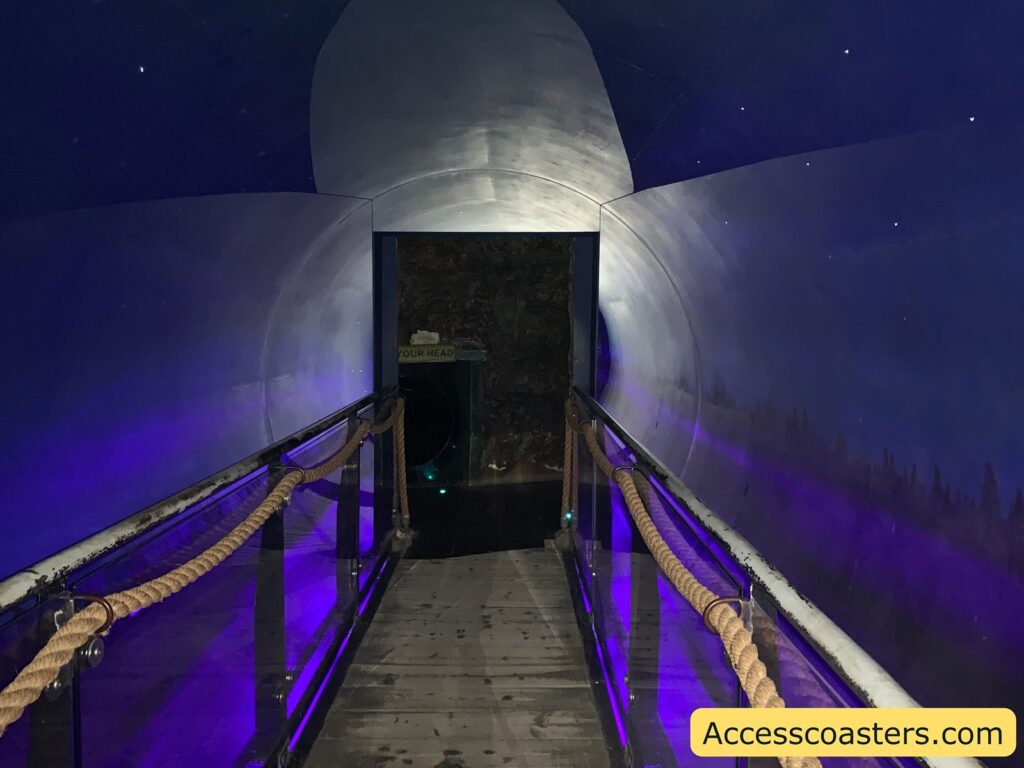 A dimly lit tunnel with a wooden walkway and rope handrails on either side. The walls and ceiling of the tunnel are painted in dark blue with small white dots resembling stars. At the end of the tunnel, there is a circular opening with a sign above it that reads "YOUR HEAD." The floor appears to be made of wooden planks. The lighting inside the tunnel has a purple hue. In the bottom right corner, there is a yellow rectangle with the text "Accesscoasters.com."