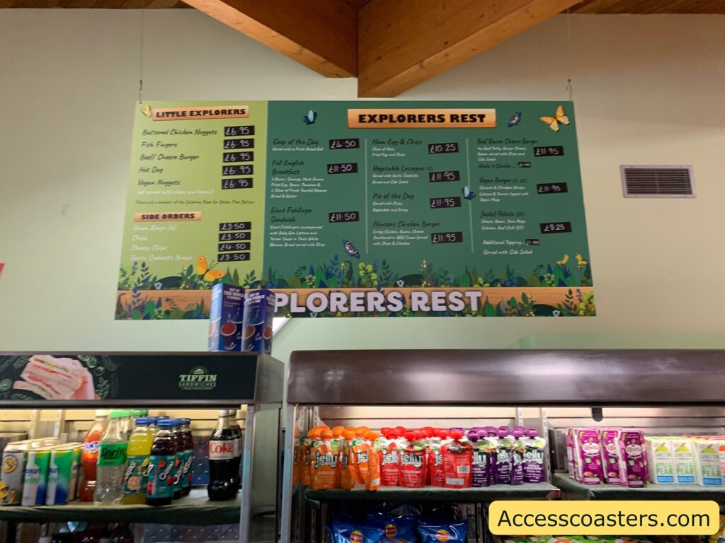 A menu board titled "Explorers Rest" is displayed, featuring two sections: "Little Explorers" and "Explorers Rest." 

Under "Little Explorers," the options include:
- Buttermilk Chicken Nuggets: £6.95
- Fish Fingers: £6.95
- Beef Cheese Burger: £6.95
- Hot Dog: £6.95
- Vegan Nuggets: £6.95

Side orders include:
- Onion Rings (6): £3.50
- Chips: £3.50
- Cheesy Chips: £4.50
- Garlic Ciabatta Bread: £3.50

Under "Explorers Rest," the options include:
- Soup of the Day: £4.50
- Full English Breakfast: £11.50
- Pie of the Day: £11.95
- Ham, Egg & Chips: £10.25
- Vegetable Lasagne: £11.95
- Hunter Chicken Burger: £11.95
- Beef Bacon Cheese Burger: £11.95
- Vegan Burger: £11.95
 Giant Fish Finger Sandwich: £11.50
- Jacket Potato: £8.25

Below the menu, there are shelves with various drinks, including bottled water, Sprite, Coca-Cola, and other beverages. There are also cartons of drinks like Ribena and other fruit-flavored drinks. The bottom right corner of the image has a watermark that reads "Accesscoasters.com."