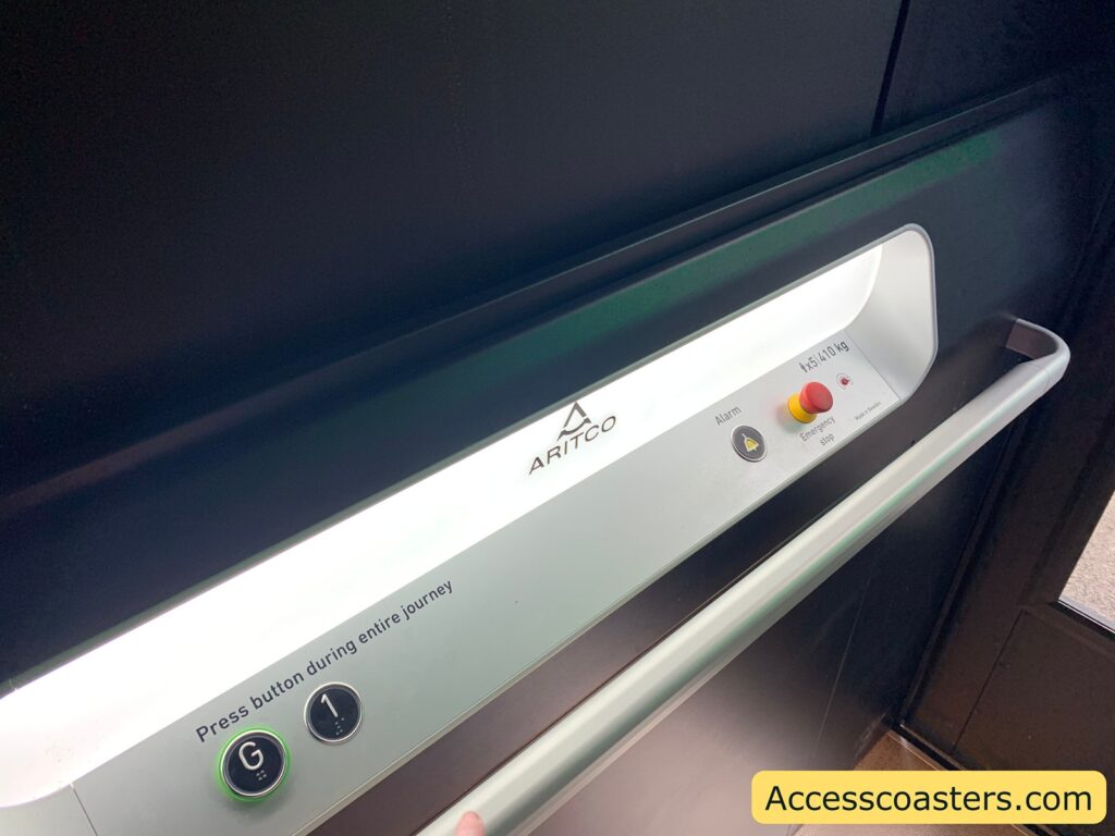 Control panel inside an elevator. The panel includes a logo for "Aritco" and a weight limit of 410 kg. There are two buttons labeled "G" and "1" with green lights around them. A message reads "Press button during entire journey." There is also an alarm button and an emergency stop button, which is red. A handrail runs along the panel. The bottom right corner has the text "Accesscoasters.com."