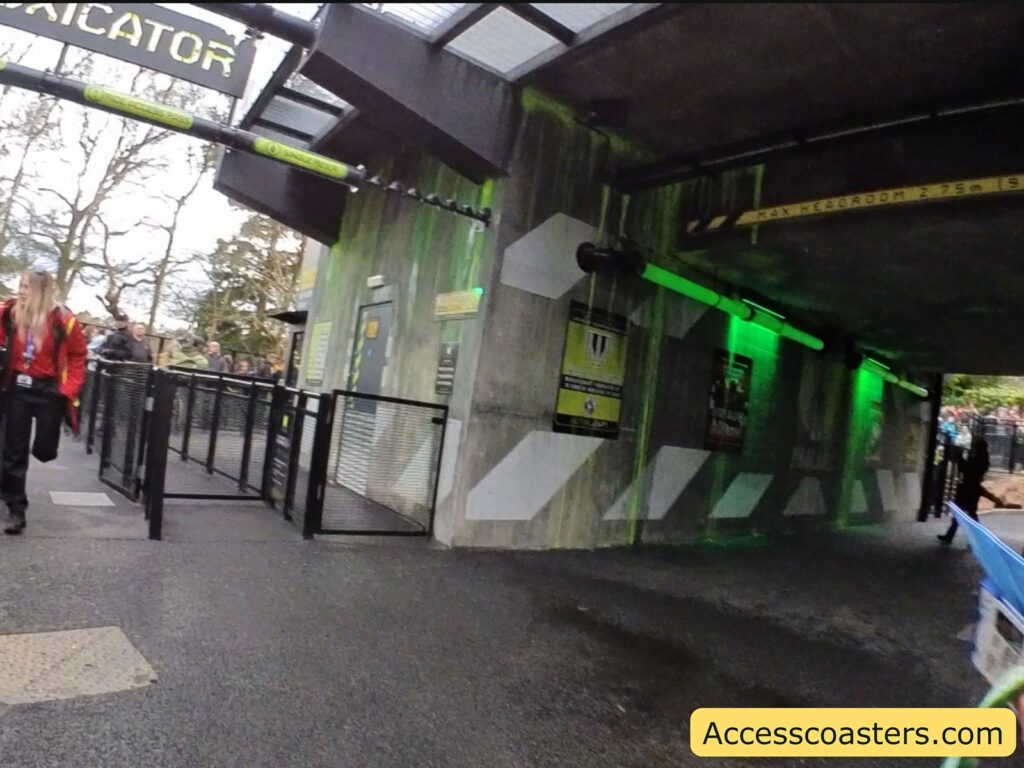 Entrance to a theme park ride with a sign above that partially reads "CATOR." The area is designed with a futuristic or industrial theme, featuring concrete walls and green lighting. A person in a red jacket is walking on the left side, and there are several people in the background. The path is lined with black metal railings, and there are posters and signs on the walls. A yellow sign above the entrance indicates "MAX HEADROOM 2.75m 9'." The website "Accesscoasters.com" is displayed in the bottom right corner.
