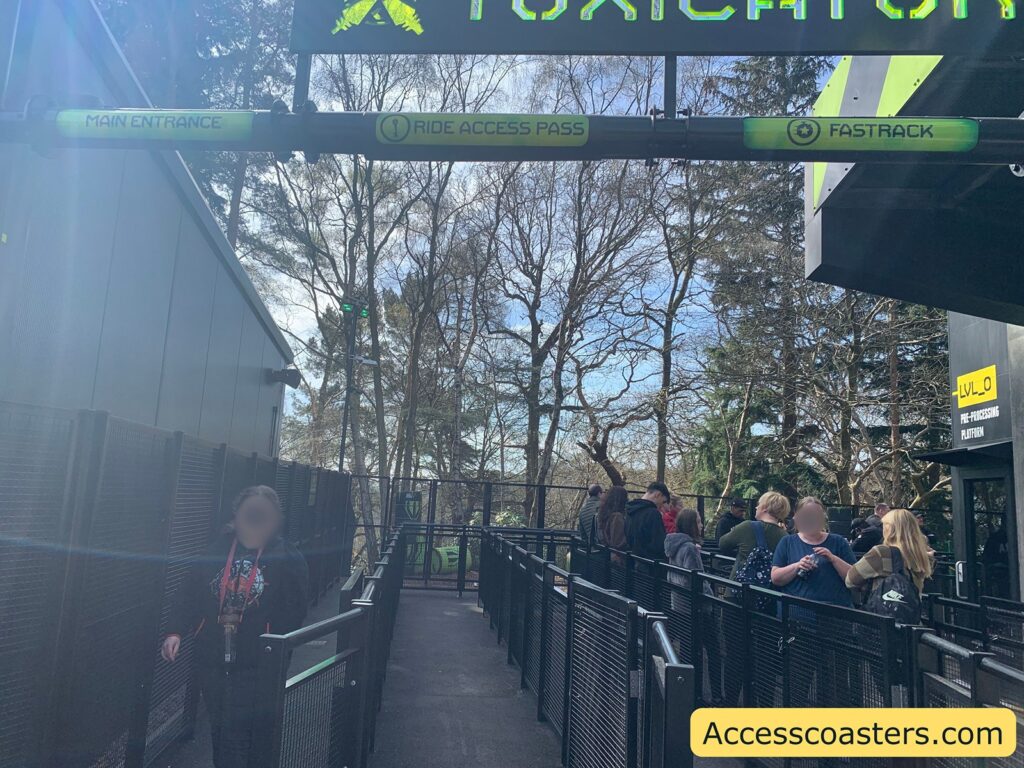 Queue area for a ride called "Toxicatron" at an amusement park. The entrance is divided into three sections: "Main Entrance," "Ride Access Pass," and "Fastrack." There are several people waiting in line, and the area is surrounded by black metal fencing. A sign on the building to the right reads "LVL_0 Pre-Processing Platform." The background features trees and a clear sky. The website "Accesscoasters.com" is displayed in the bottom right corner.