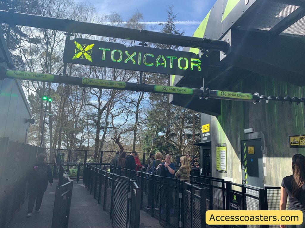 Entrance to a theme park ride called "TOXICATOR." The sign is in bright green letters with a toxic symbol. There are several lanes for entry, labeled "Main Entrance," "Ride Access Pass," "Fastrack," and "Single Rider." People are queuing in the lanes, and the area is surrounded by black metal railings. The building has a grey and green color scheme with a sign indicating "LVL_0" and "Platform." There are warning signs about restricted access and speed zones. The sky is clear with some trees in the background. A website URL, "Accesscoasters.com," is displayed at the bottom right corner.