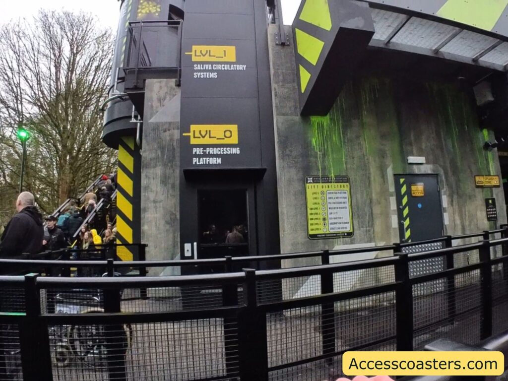 a lift with a futuristic and industrial design. The structure has a dark color scheme with yellow accents and signage. There are two levels marked as "LVL_1" and "LVL_0." "LVL_1" is labeled as "SALIVA CIRCULATORY SYSTEMS" and "LVL_0" as "PRE-PROCESSING PLATFORM." A group of people is seen ascending a staircase on the left side of the building. The exterior wall has green streaks, possibly simulating some kind of fluid or slime. There is a door with a sign that reads "STAFF ACCESS." A signboard lists various levels and their corresponding functions. The website "Accesscoasters.com" is displayed at the bottom right corner.