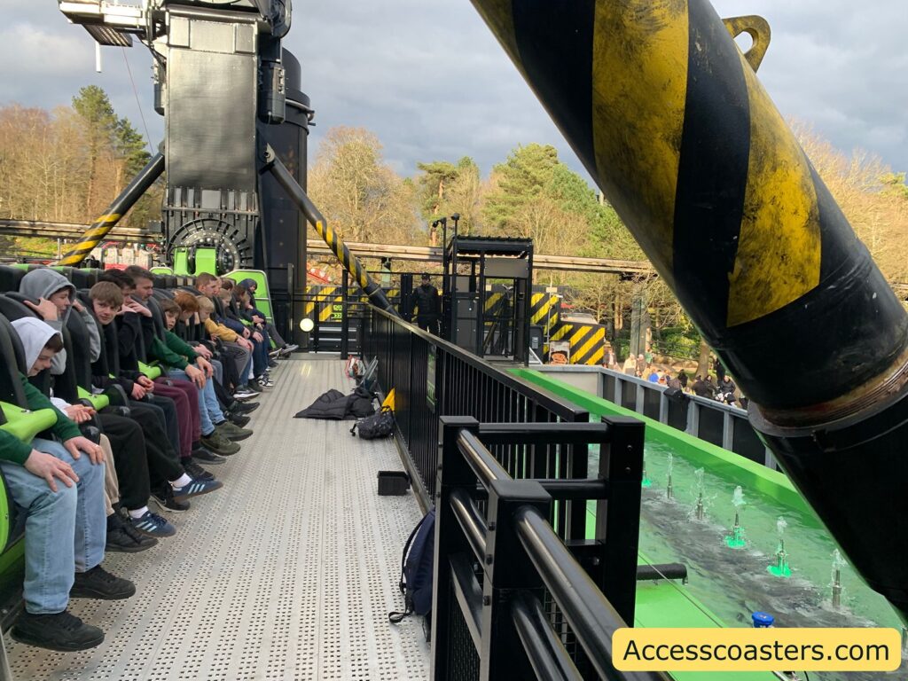 A group of people is seated in a row on a theme park ride platform, likely waiting for the ride to start. They are sitting in seats with green and black restraints. The platform is metallic with a perforated surface. In the background, there are trees and a cloudy sky. A large black and yellow striped structure is visible, possibly part of the ride's mechanism. On the right side, there is a green trough with water fountains. The website "Accesscoasters.com" is displayed in the bottom right corner.