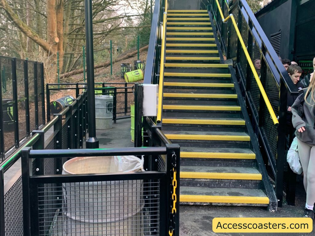 A set of outdoor stairs with yellow edges and black railings. To the left of the stairs, there is a path lined with black mesh fencing and a few metal barrels, some of which have green and black designs. A group of people is visible on the right side, near the bottom of the stairs. The background features trees and a wooded area. In the bottom right corner, there is a yellow box with the text "Accesscoasters.com".