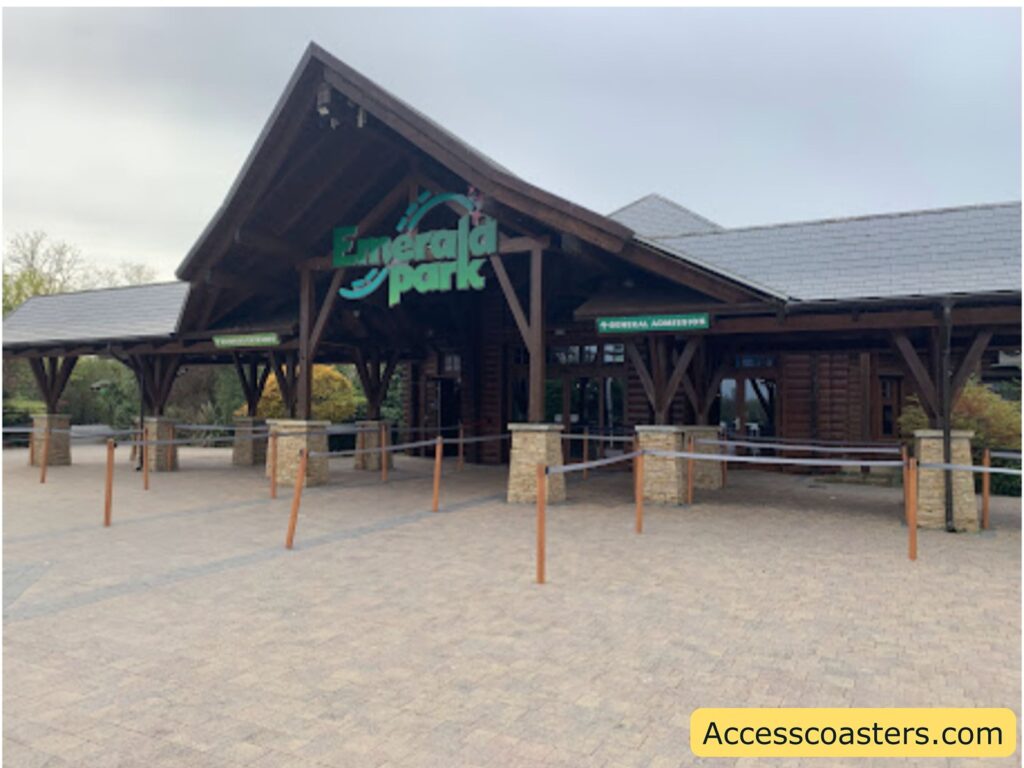 Wooden entrance building with a peaked roof The sign above the entrance reads "Emerald Park" in large green letters, with a stylized roller coaster track graphic. There are two smaller signs: one on the left says "TICKETS/ONLINE ENTRY" and one on the right says "GENERAL ADMISSION." In front of the entrance, there are queue barriers made of wooden posts and ropes. The ground is paved with light brown bricks, and there are some bushes and trees in the background.