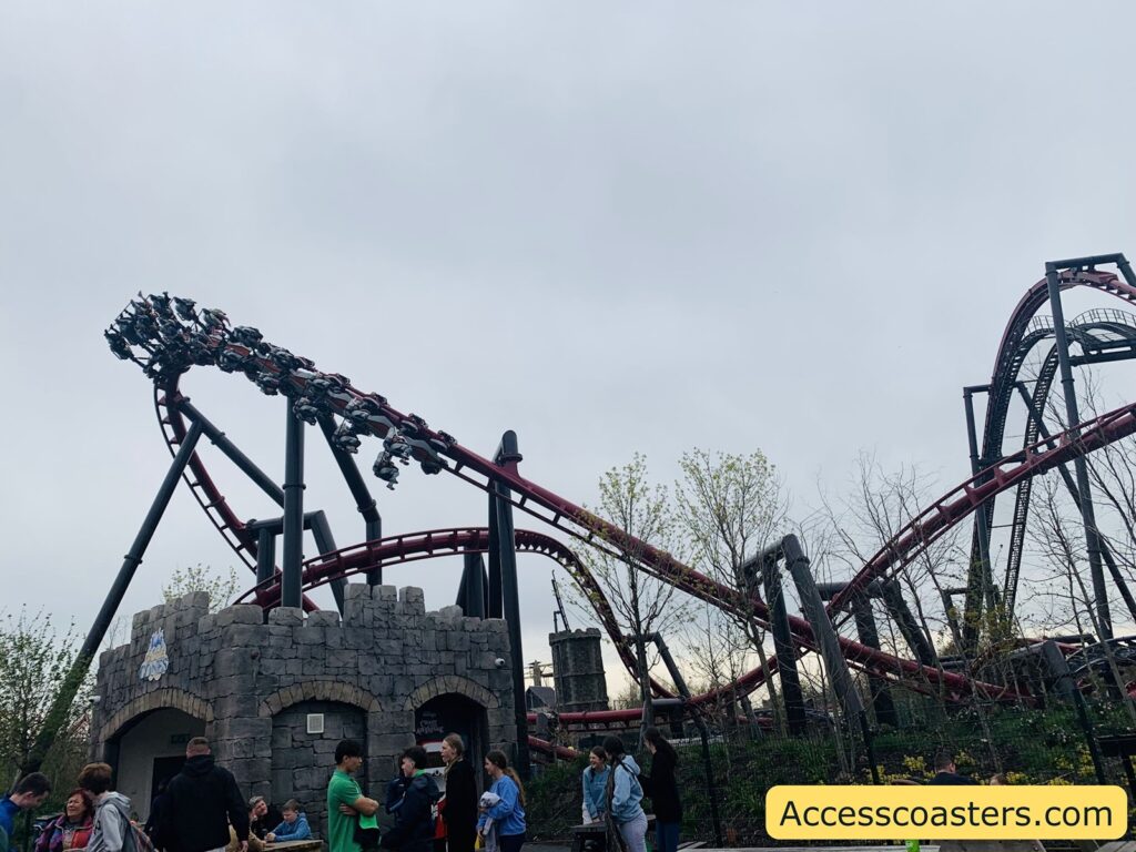 Outdoor amusement park scene featuring a large, looping roller coaster with red tracks and black supports. The coaster train is visible on the upper left, filled with riders, some with their legs dangling as it’s an inverted coaster. Below the coaster, there is a stone building resembling a medieval castle, with people gathered around the entrance and queuing. 