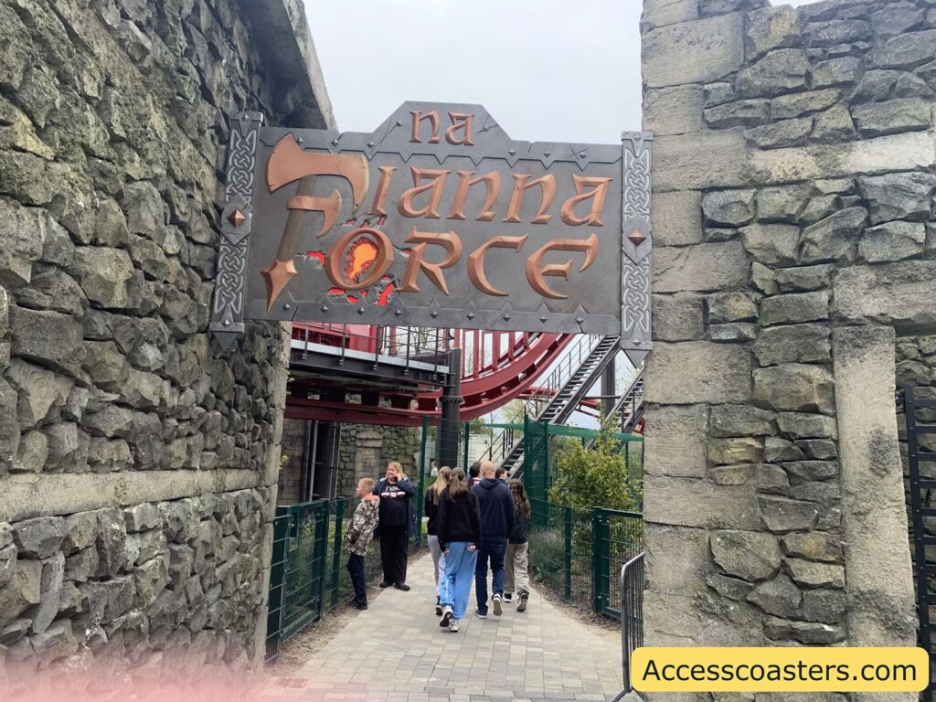 Stone archway with a sign reading "Na Fianna Force" in stylized lettering, featuring Celtic knot designs on the sides. The sign hangs between two stone walls, resembling a castle or fortress entrance. Below the sign, a group of people, including adults and children, are walking along a paved path with a metal fence on one side. In the background, you can see part of a red roller coaster track