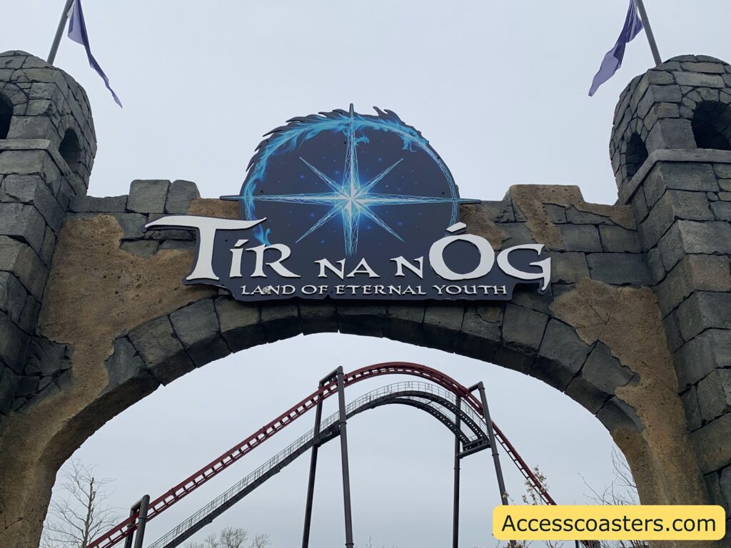 Stone archway entrance with two small castle-like towers on each side. At the top center of the arch is a large sign that reads: "Tír na nÓg LAND OF ETERNAL YOUTH" in stylized white letters on a dark background with a glowing blue starburst design. Behind the arch, part of a red roller coaster track is visible, curving upwards.