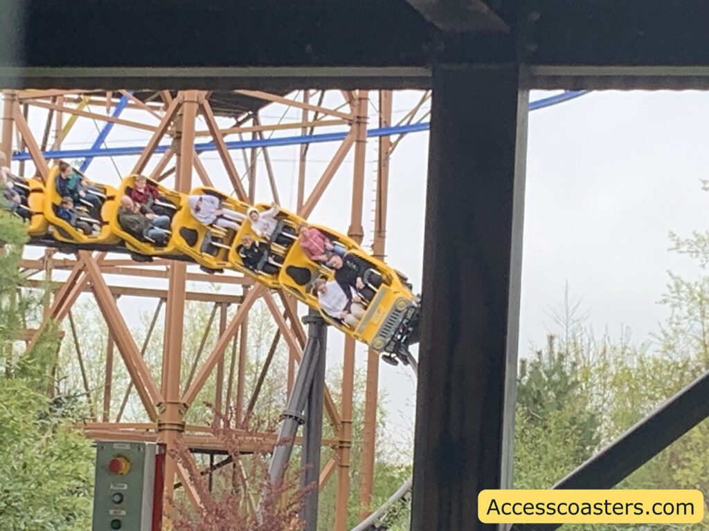 Roller coaster train with yellow cars, filled with people, curving sharply on a steel track. The riders appear to be enjoying the thrill, with some raising their hands and others gripping the safety bars. The coaster cars have a black spotted pattern, resembling a cheetah or animal print. 