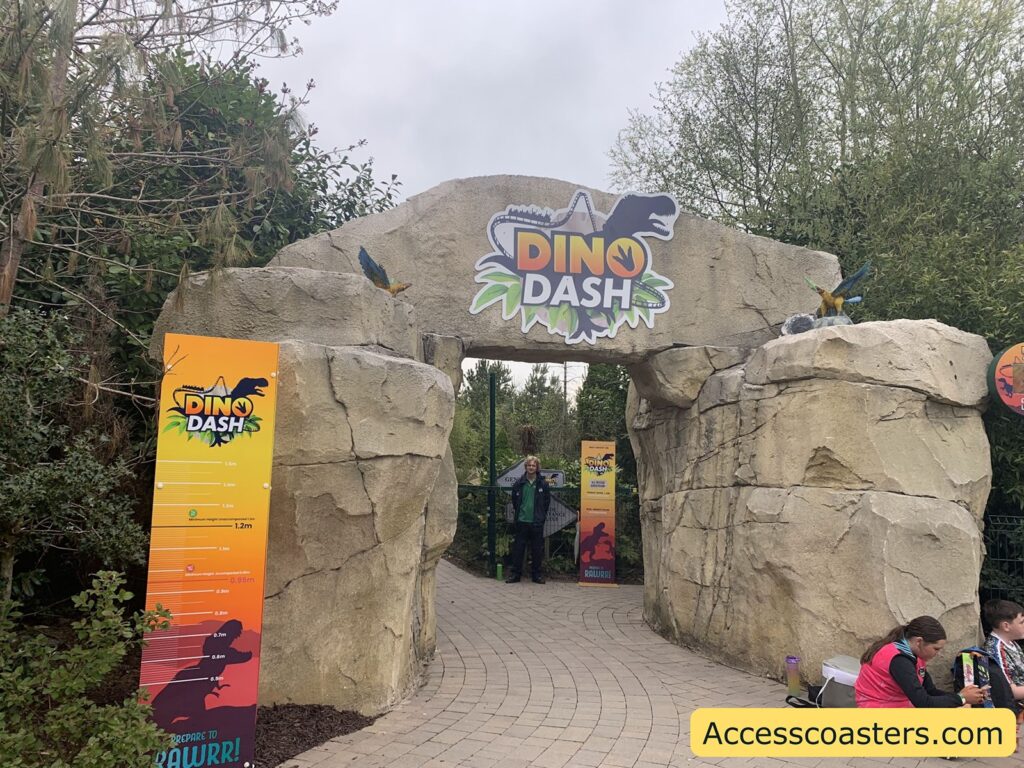 tone archway entrance to a theme park ride called "Dino Dash." The archway is made to look like large, rough rocks with two small dinosaur models perched on top. Above the arch is a colorful sign with the ride's name, featuring a dinosaur silhouette and leafy graphics. On the left side, there is a tall, gradient-colored height measurement board with the Dino Dash logo, showing various height requirements for the ride. Two staff members in green jackets stand under the arch, and two people are sitting on the ground to the right, near the rocks. 