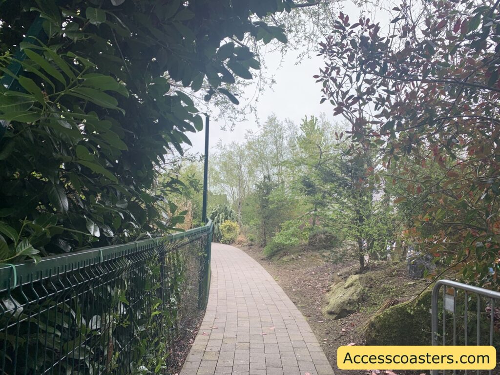 A winding paved pathway bordered by a green metal fence on the left and various trees and bushes on both sides. The path curves gently to the right